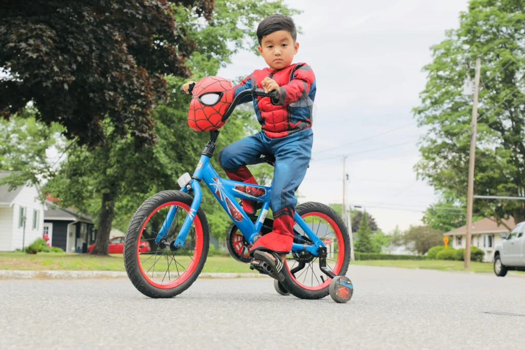 Elementary Age boy wearing Spider-Man costume riding a red and blue bike with training wheels